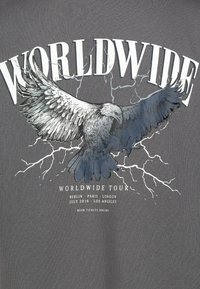 Graues T-Shirt mit einer detaillierten Illustration eines Adlers mit ausgebreiteten Flügeln, Blitzen und Text für eine Welt-Tour.