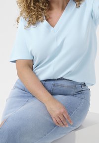 Lyseblå v-hals top med korte ærmer, glat tekstur. Sammen med lyseblå denimnederdel med slids. Hånd hvilende på lår.