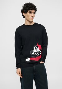 Νεαρός άνδρας με σγουρά μαλλιά που φοράει μαύρο πουκάμισο με μακριά μανίκια, με μεγάλο κόκκινο, λευκό και μαύρο σχέδιο καρτούν αλεπούς, σε συνδυασμό με σκούρο τζιν.