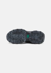 Suela de zapato de goma negra con profundos surcos y un patrón texturizado. Presenta un logo verde en el centro y una entresuela negra suave.