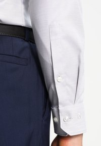 Chemise habillée à motif gris clair avec des poignets boutonnés, assortie à un pantalon bleu foncé. Accent sur le tissu en coton et la couture détaillée.