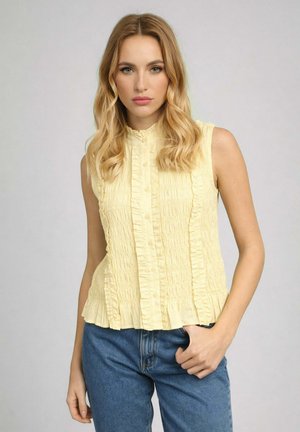 Femme blonde aux cheveux ondulés lâchés, portant un chemisier sans manches jaune pâle à volants et un jean bleu, debout devant un fond clair uni.