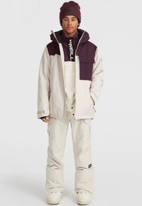 Beige snow jacket with a purple upper panel, zippered pockets, paired with beige snow pants and white boots, worn with a dark beanie.