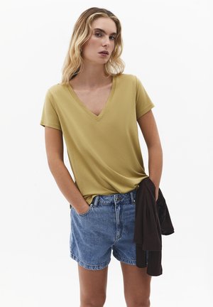 Jonge vrouw met blond haar, gekleed in een mosterdgele T-shirt met V-hals en denim shorts, die een zwart kledingstuk in haar linkerhand vasthoudt.