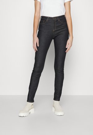 Slim-fit mørkeblå jeans båret med hvide chunky støvler og en simpel hvid t-shirt med korte ærmer mod en lys baggrund.