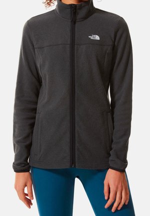 Osoba ubrana w ciemnoszarą polarową kurtkę North Face na zamek błyskawiczny z długimi rękawami oraz niebieskie legginsy, stojąca na białym tle.