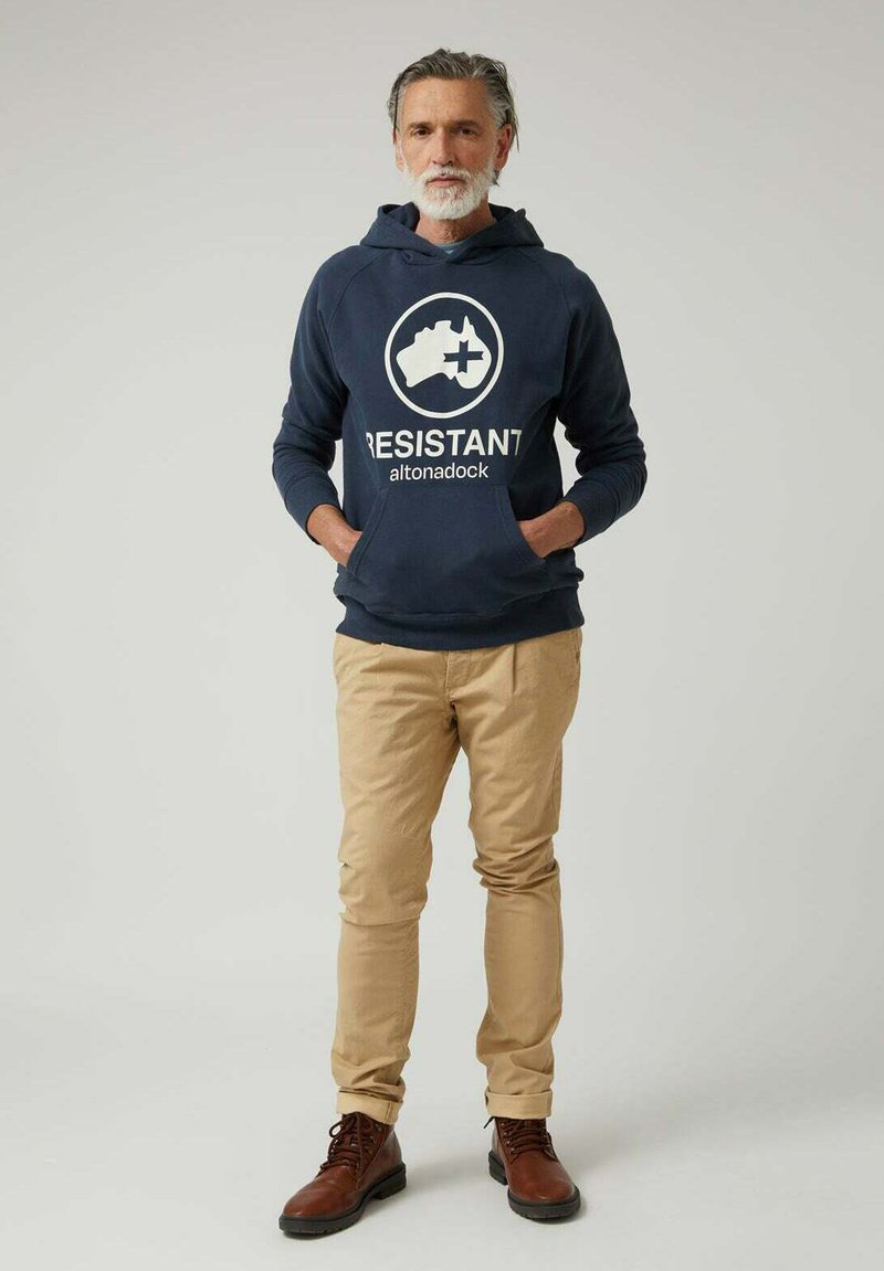 Sudadera azul marino con el logo "RESISTANT", bolsillo frontal, combinada con pantalones beige y botas marrones. Estilo casual y ajuste moderno.
