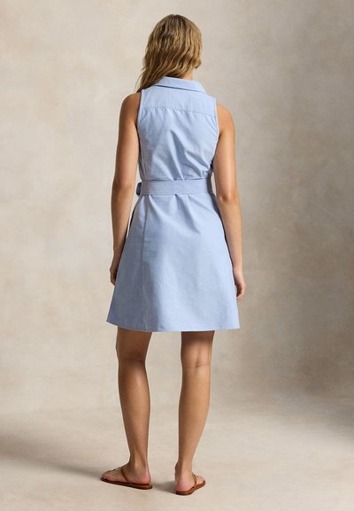 Femeie purtând o rochie albastru deschis fără mâneci, cu centură, și sandale plate maro, stând cu spatele către privitor, pe un fundal neutru.