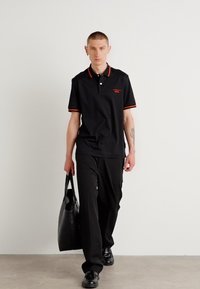 Polo noir avec col et accents rouges, associé à un pantalon et des chaussures noirs. Portant un grand sac fourre-tout noir. Look simple et moderne.