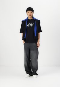 Black t-shirt with white graphic, paired with baggy gray jeans and white sneakers. Blue-accented jacket draped over shoulders.