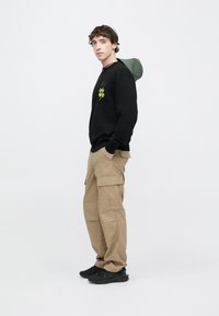 Felpa nera con cappuccio verde e grafica di trifoglio, abbinata a pantaloni cargo beige e sneakers nere. Stile streetwear semplice e funzionale.