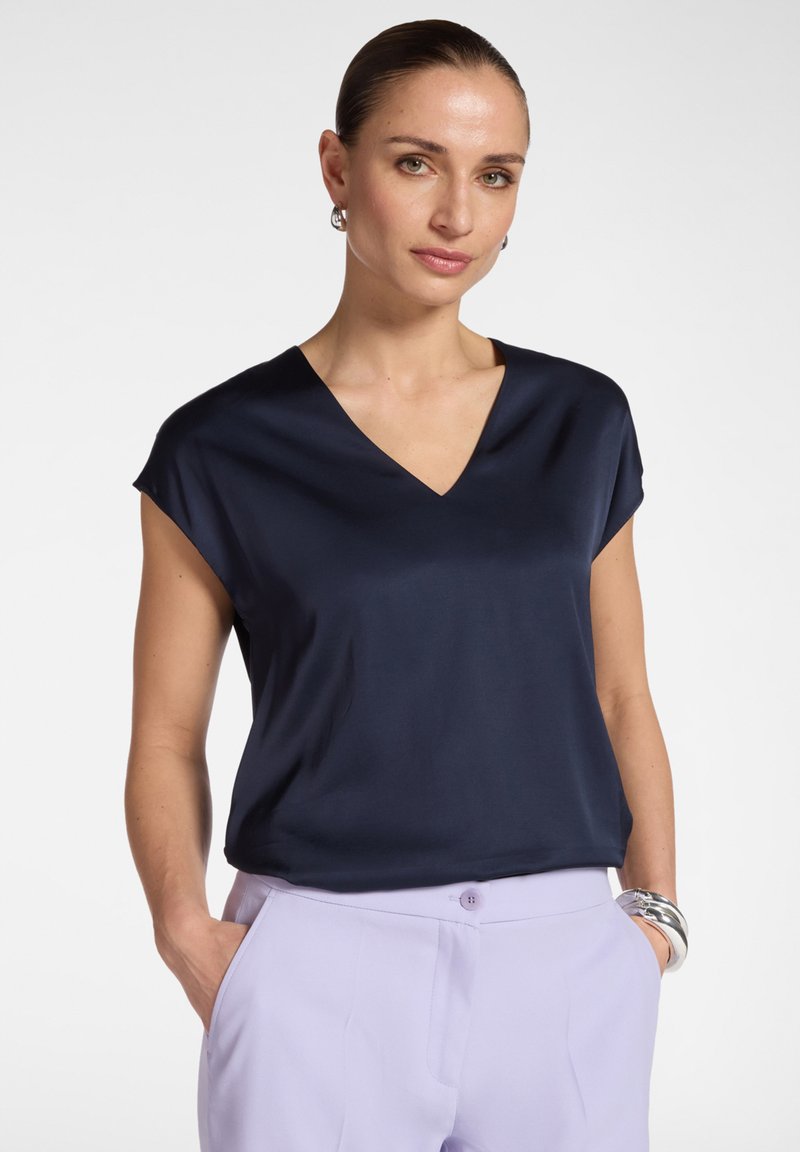 Mujer con blusa azul marino de escote en V y pantalones lavanda claros, manos en los bolsillos, pulsera de plata y pendientes de aro, sobre un fondo blanco.