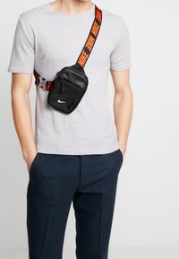 Černá crossbody taška Nike s červeným popruhem s logem "Nike", s texturovaným povrchem, zipovým uzávěrem a logem. Nositá přes šedé tričko.