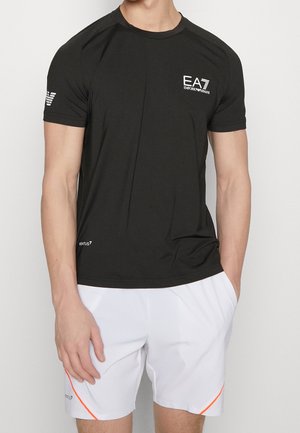 Mann trägt ein schwarzes EA7 Emporio Armani Kurzarmshirt und weiße Sportshorts mit orangefarbenen Akzenten, steht vor einem einfarbigen Hintergrund.