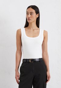 Witte mouwloze top met een ronde hals, gemaakt van zachte stof. Gecombineerd met zwarte broeken en een riem met een gouden gesp.