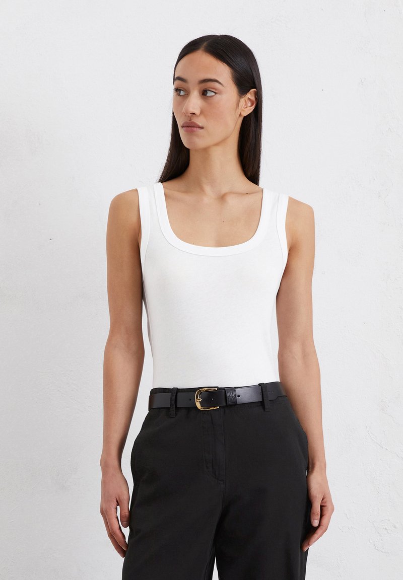 Witte mouwloze top met een ronde hals, gemaakt van zachte stof. Gecombineerd met zwarte broeken en een riem met een gouden gesp.