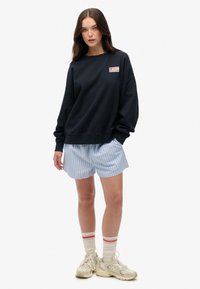Navy sweatshirt with a small flag patch, paired with light blue striped shorts. Model wears beige sneakers and red-accented socks.
