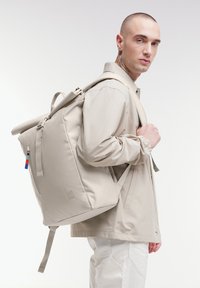 Sac à dos beige à rabat avec une texture lisse, des sangles ajustables, une poche latérale zippée et un détail de logo. Présenté sur un modèle portant une veste légère.