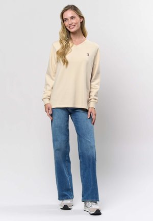 Beige V-hals sweatshirt met lange mouwen, voorzien van een geborduurd logo. Gecombineerd met lichtblauwe wijde spijkerbroek en grijze sneakers.