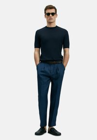 Marine ribbed short-sleeve trui, blauwe geplooide broek en zwarte sandalen. Model staat met handen in de zakken tegen een witte achtergrond.