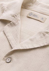 Veste en tissu texturé couleur crème avec col à revers, fermeture par un seul bouton, et une étiquette Ombre Heritage taille medium à l'intérieur.