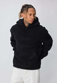 Hoodie en polaire noire avec une fermeture éclair partielle, une poche kangourou et un tissu texturé. Présente une coupe décontractée et des poignets côtelés.