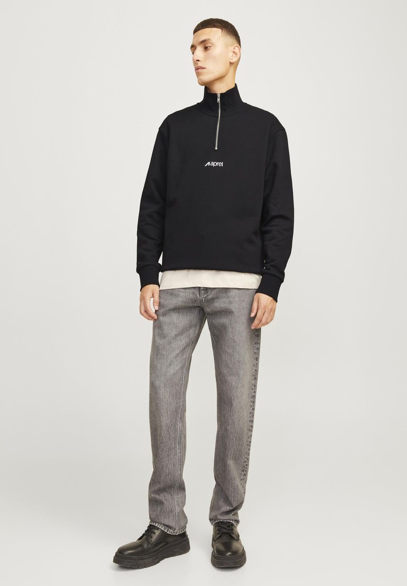 Sweat-shirt noir à fermeture éclair avec un col montant et un logo blanc, associé à un jean gris et des chaussures noires. Le tissu semble lisse et ajusté.