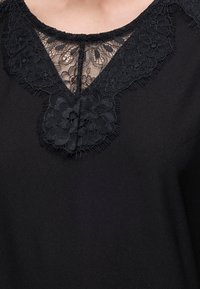 Haut noir avec des détails en dentelle florale autour de l'encolure et une insertion en dentelle transparente au niveau de la poitrine.