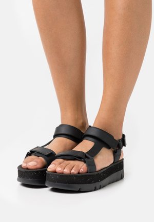 Black platform sandals with adjustable straps, made of smooth synthetic material. Features a speckled rubber sole for textured grip.