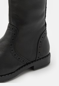 Bottine en cuir noir avec une texture lisse, dotée d'accents perforés et d'un petit talon carré, conçue pour le confort et le style.