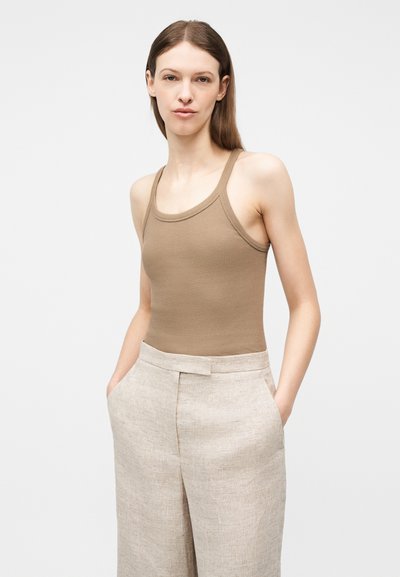 Femme aux cheveux bruns et lisses portant un débardeur beige et un pantalon large en lin clair, les mains dans les poches, sur un arrière-plan uni clair.