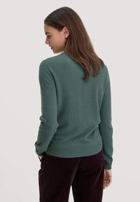 Teeingestrickter Pullover in Tealtönen mit rundem Ausschnitt und geripptem Saum. Das Modell hat lange Haare und trägt dunkelburgunderfarbene Cordhosen.