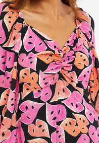 Czarna bluzka z tkaniny z liściastym wzorem w odcieniach różu i pomarańczy, z marszczonym środkiem i dekoltem w kształcie litery V. Krótkie rękawy z delikatnym rozkloszowaniem.
