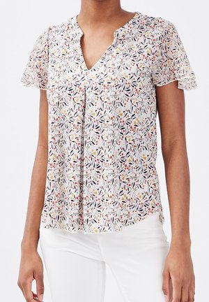 Femme portant un chemisier blanc à fleurs à manches courtes volantées et un pantalon blanc, posant devant un fond uni.