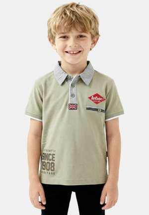 Jeune garçon aux cheveux blonds souriant, portant un polo vert clair avec un col gris et un texte imprimé "Depuis 1908" sur le devant.