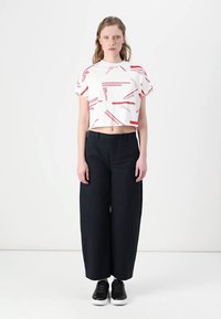Krátky biely tričko s červeným grafickým textom, v kombinácii s voľnými čiernymi nohavicami. Model má na sebe čierne topánky. Jednoduché pozadie.