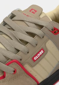 Baskets Globe beige et sable avec des lacets beige épais, des écussons de logo rouges, des œillets rouges et une semelle noire visible.