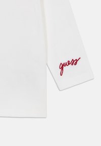 Hvit langermet t-skjorte med en rød brodert "guess"-logo ved mansjetten. Glatt stoff med en avslappet passform.