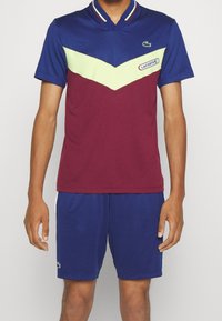 Lacoste polo- och shorts-set i blått, vinrött och limegrönt. Har en färgblockeringsdesign med textilstruktur och en krage. Logotypdetalj förekommer.