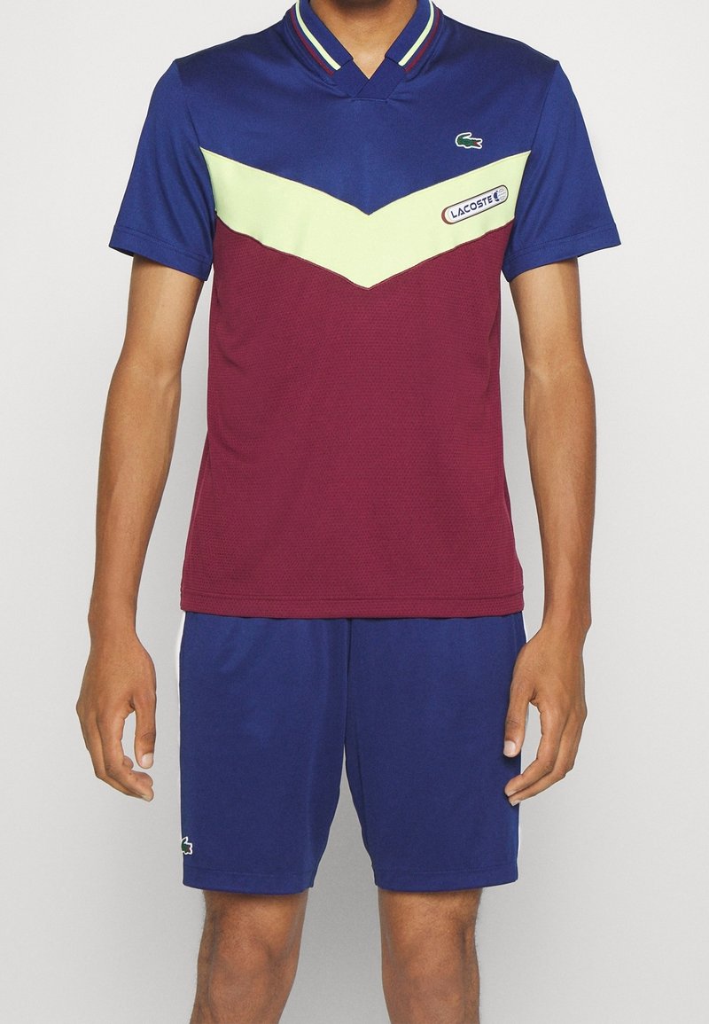 Lacoste polo- och shorts-set i blått, vinrött och limegrönt. Har en färgblockeringsdesign med textilstruktur och en krage. Logotypdetalj förekommer.