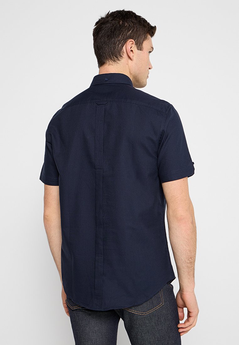 Homme portant une chemise à manches courtes boutonnée bleu marine foncé et un jean sombre, vu de dos sur un fond blanc uni.