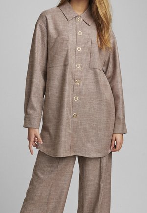 Femme portant une chemise boutonnée marron à carreaux amples avec un pantalon large assorti, debout devant un fond gris clair uni.