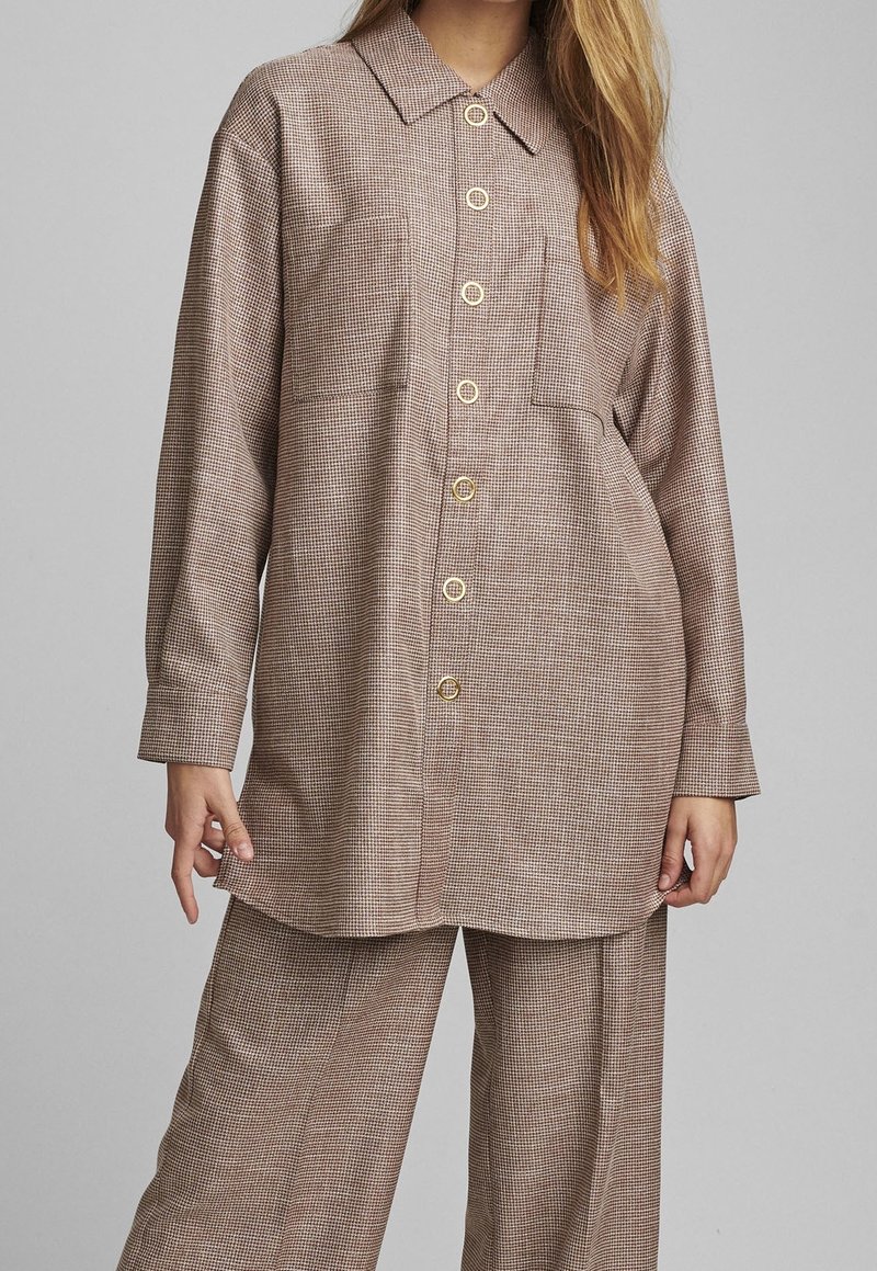 Femme portant une chemise boutonnée marron à carreaux amples avec un pantalon large assorti, debout devant un fond gris clair uni.