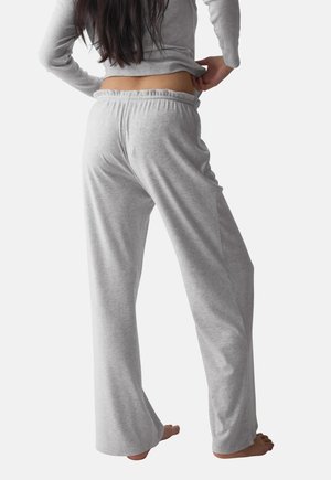 Femme aux longs cheveux foncés portant un pantalon et un haut de détente gris clair, debout pieds nus, mains ajustant la taille, de dos.