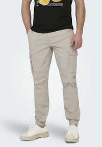 Pantalones cargo beige con puños elásticos, múltiples bolsillos laterales y un corte recto. Combinados con zapatillas blancas y una camiseta gráfica negra.