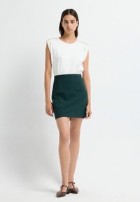 Top bianco senza maniche con decorazioni sulle spalle, abbinato a una gonna verde aderente e scarpe con cinturino marroni, su uno sfondo semplice.
