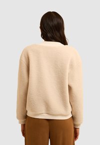 Felpa beige morbida con vestibilità rilassata, colletto e polsini a coste e superficie texturizzata. Indossata con pantaloni marroni, vista da dietro.