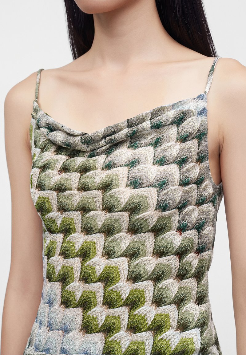 Femme portant un haut sans manches avec un motif tissé texturé et multicolore dans des tons de vert, beige et bleu.