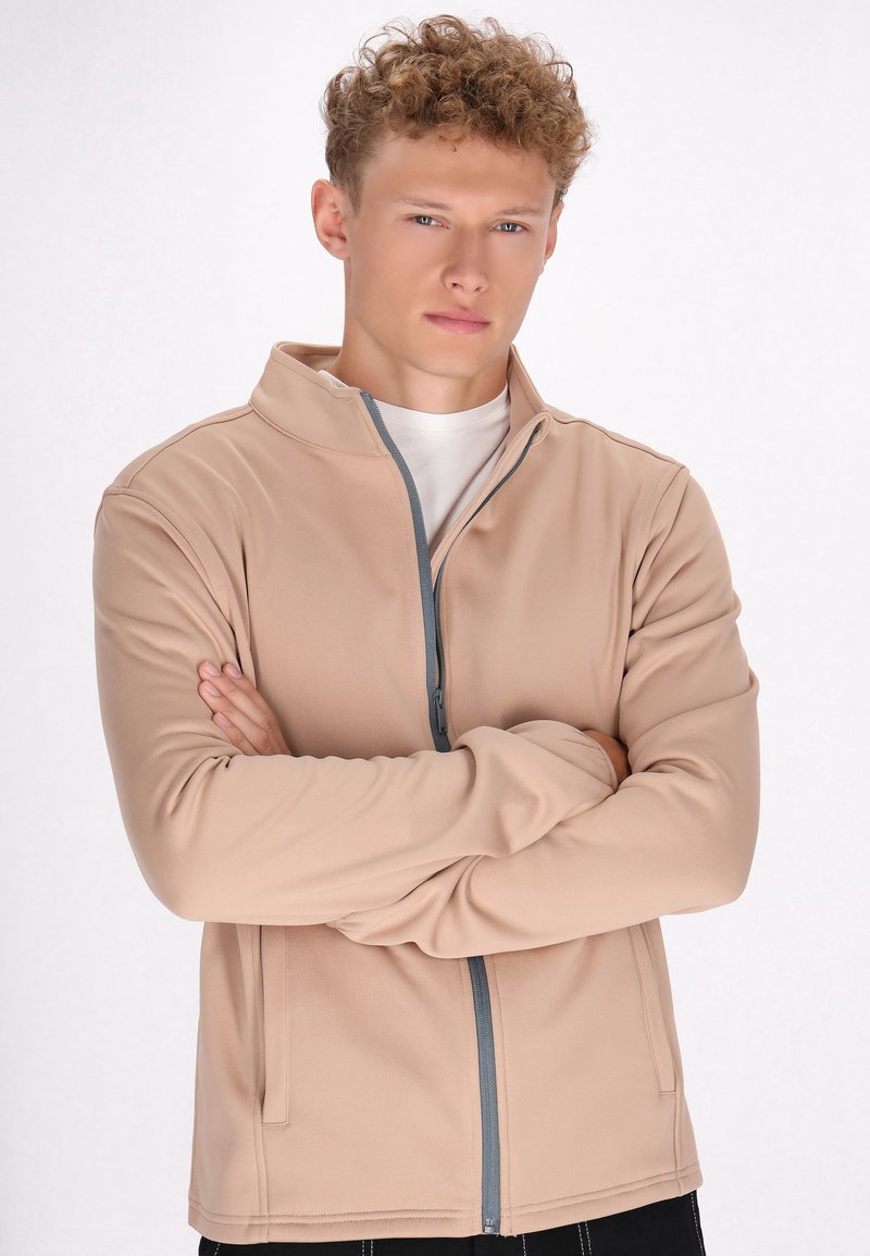 Beige ritsjack gemaakt van zachte stof met een opstaande kraag en grijze ritsdetails; model heeft de armen gekruist tegen een neutrale achtergrond.