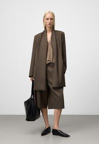 Frau in übergroßem braunen Blazer und knielangen Shorts, die eine schwarze Handtasche hält und auf einem grauen Boden vor einer weißen Wand steht.